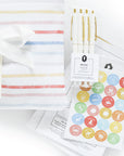Detailed view of the 'Bundle of Joy' gift set from The Everyday Mother, showing additional contents such as white and gold pens, milestone stickers, and doctor visit note sheets. These accessories are designed to enhance the experience of tracking baby's milestones and daily routines, presented in a clear plastic sleeve beside the striped journal package with a white satin ribbon.