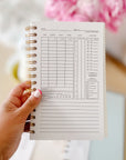 A hand holding an open page of The Everyday Mother daily baby tracker, which features detailed sections for documenting nursing, feeding, pumping, sleep, and diaper changes. The page layout is clean and structured, with checkboxes for reminders and a notes section at the bottom. Soft pink flowers in the blurred background add a gentle, aesthetic touch, enhancing the focus on the journal in a nurturing and organized setting.