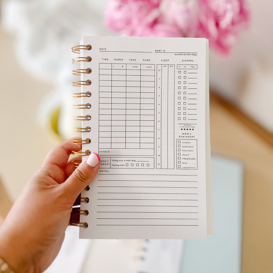 A person holding an open Daily Baby Tracker from The Everyday Mother with an undated weekly schedule layout, featuring sections for the days of the week, a breastfeeding log, and a notes area.