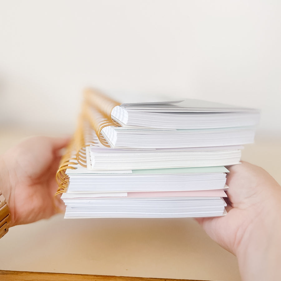 A stack of spiral notebooks, including The Everyday Mother's Daily Baby Tracker, held in a hand against a neutral background.