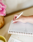 A hand writing in a spiral-bound Everyday Mother daily baby tracking journal with a white pen. The journal has sections for time, nursing, feeding, pumping, sleep, and diapers. The background features a wooden vase with pink flowers and a green mug.