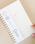 Close-up view of an open The Everyday Mother baby tracking journal. The left side shows the cover page with the brand's logo, while the right side features the title '6 Month Baby Tracker' in elegant typography. The journal is designed for detailed recording of daily activities such as feedings, diaper changes, sleep, and milestones. A hand with pink manicured nails is holding the journal open, emphasizing its user-friendly design