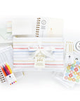 Bundle of Joy baby journal gift set includes a striped cloth with ribbon, baby trackers, colorful pens, thank you cards, milestone stickers, a 'Breathe Mama' tin, and a pack of pens. The set is designed for tracking baby's milestones and activities.