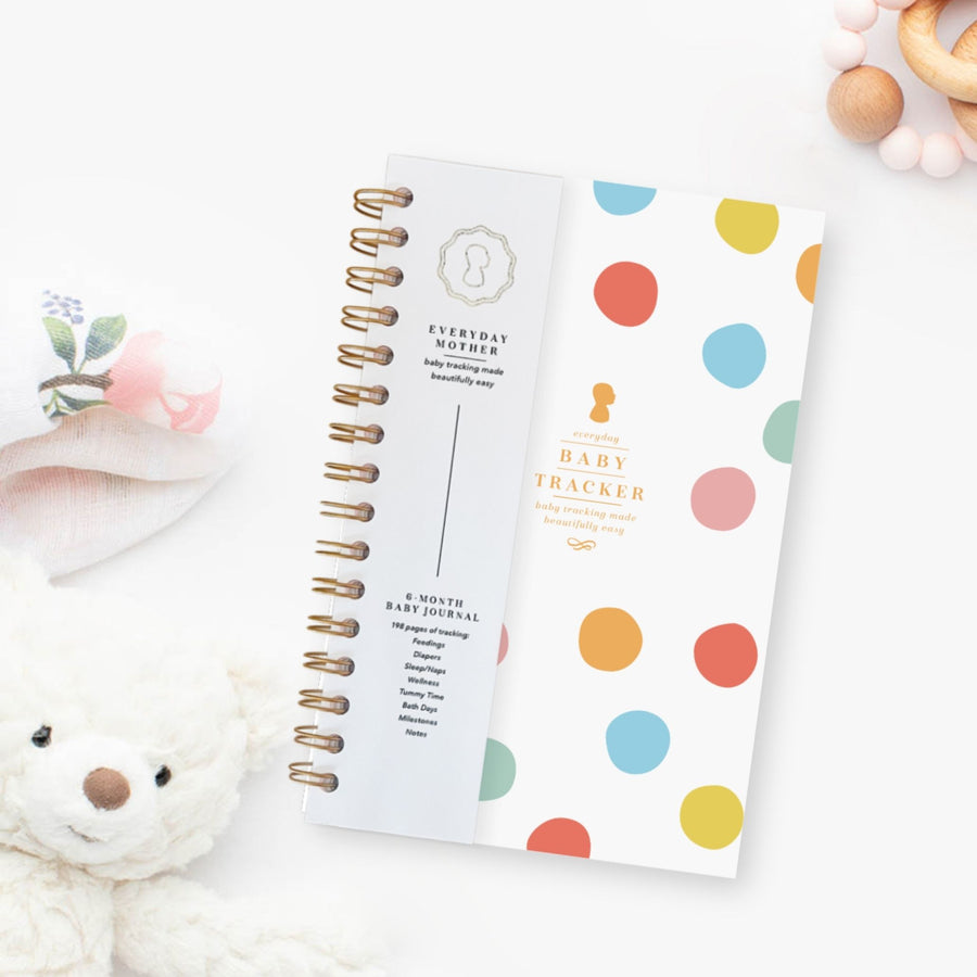 Everyday Mother baby tracker journal with gold spiral binding. Tracks 6 months of feedings, sleep, diapers, tummy time, bath days, wellness, and milestones. Background includes a plush teddy bear and a soft floral cloth.