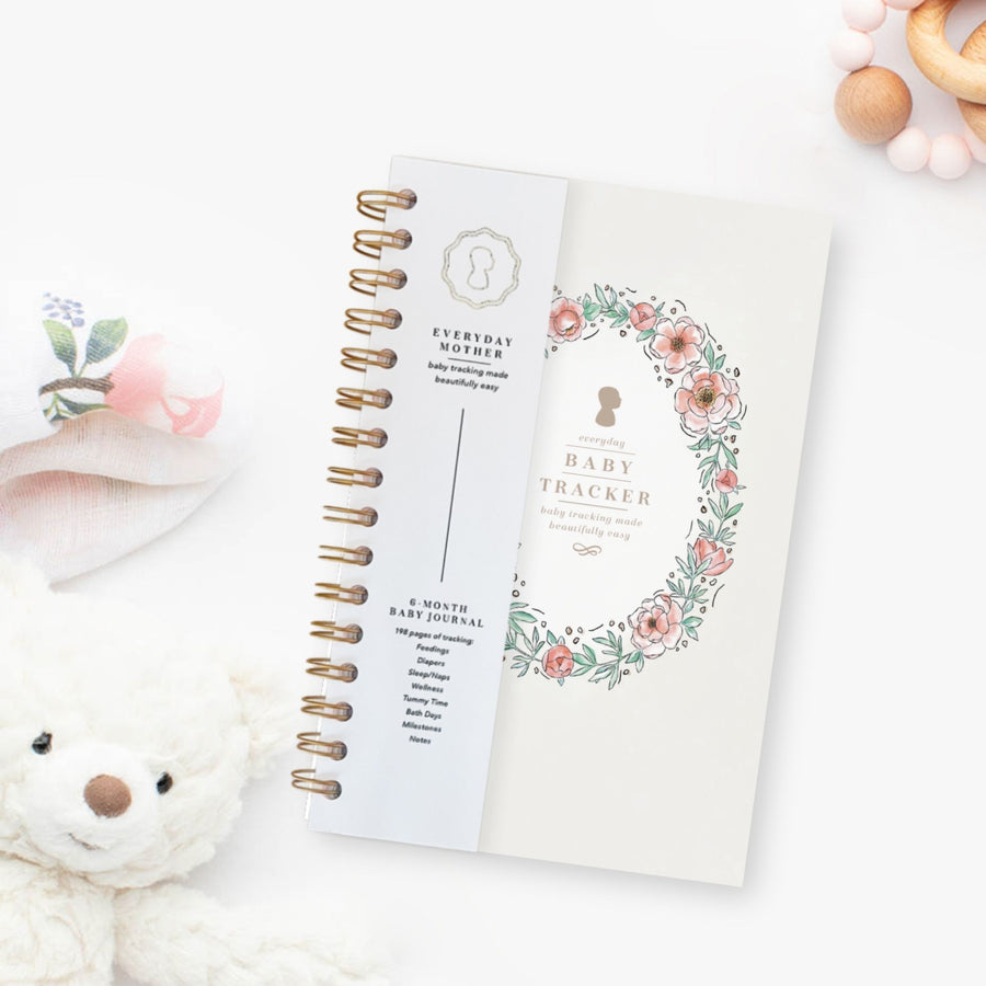 Everyday Mother baby tracker journal with gold spiral binding. Tracks 6 months of feedings, sleep, diapers, tummy time, bath days, wellness, and milestones. Background includes a plush teddy bear and a soft floral cloth.