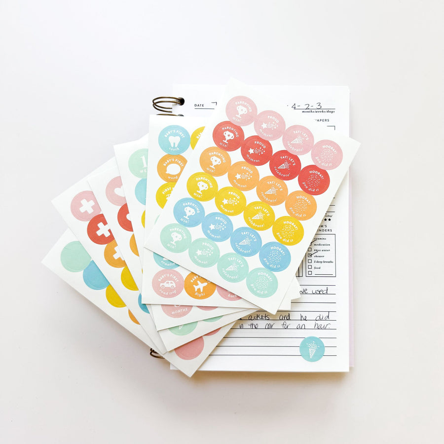 A stack of colorful The Everyday Mother Milestone Stickers with various symbols and icons attached to a clipboard on a white surface.