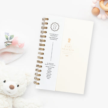 Everyday Mother baby tracker journal with gold spiral binding. Tracks 6 months of feedings, sleep, diapers, tummy time, bath days, wellness, and milestones. Background includes a plush teddy bear and a soft floral cloth.