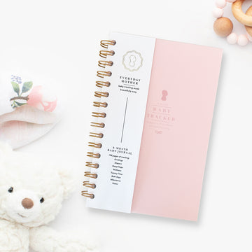 Image of the 'Everyday Mother' baby tracking journal with a light pink cover and gold spiral binding. The journal is placed on a white surface with baby items around it, including a stuffed teddy bear, a floral cloth, and a wooden teething ring. The journal tracks various baby activities like feeding, diapers, sleep, wellness, tummy time, bath days, and milestones.