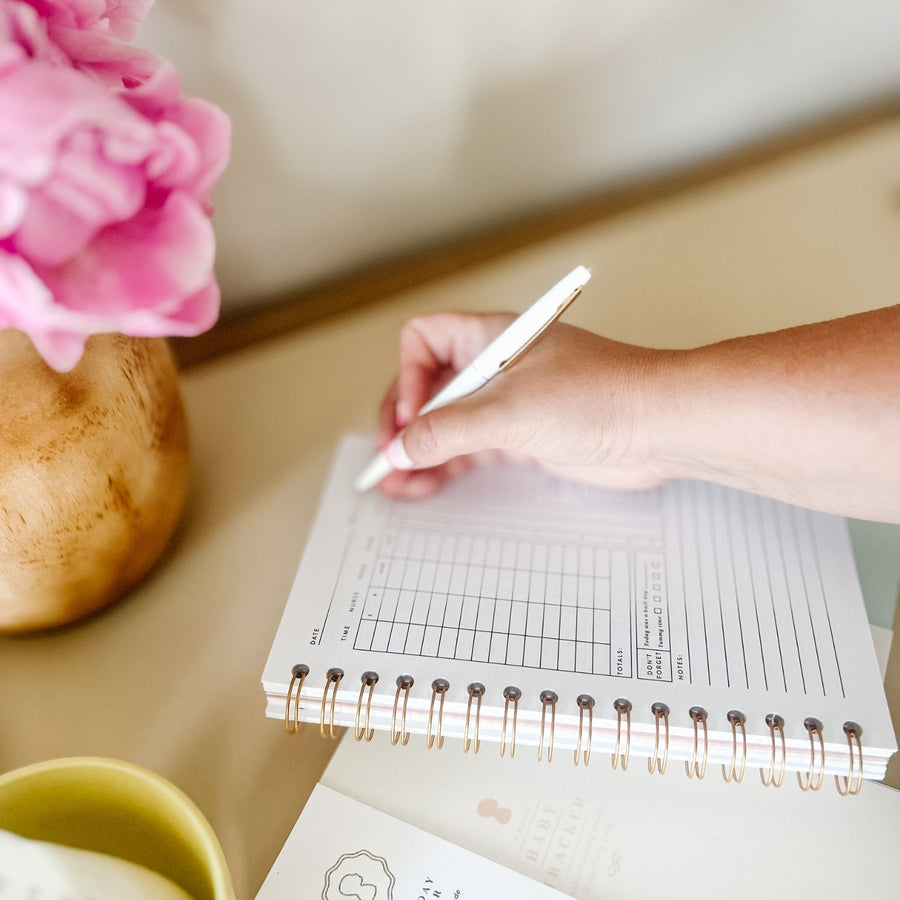 A hand writing in a spiral-bound Everyday Mother daily baby tracking journal with a white pen. The journal has sections for time, nursing, feeding, pumping, sleep, and diapers. The background features a wooden vase with pink flowers and a green mug.