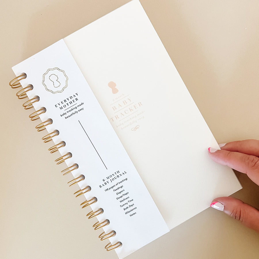 Close-up view of an open The Everyday Mother baby tracking journal. The left side shows the cover page with the brand's logo, while the right side features the title '6 Month Baby Tracker' in elegant typography. The journal is designed for detailed recording of daily activities such as feedings, diaper changes, sleep, and milestones. A hand with pink manicured nails is holding the journal open, emphasizing its user-friendly design