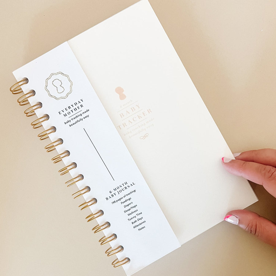 A hand touching an ivory Everyday Mother Baby Tracker book showing the ivory cover, white cover slip with gold logo, and gold spiral rings 