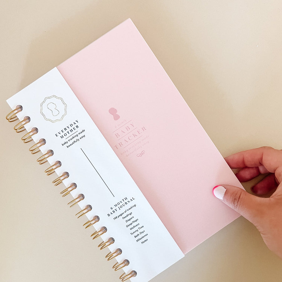 A hand touching a light pink Everyday Mother Baby Tracker book showing the ivory cover, white cover slip with gold logo, and gold spiral rings