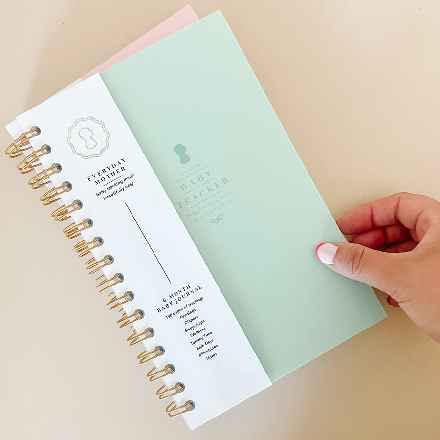 A hand touching the corner of a mint green Everyday Mother Baby Tracker book showing the ivory cover, white cover slip with gold logo, and gold spiral rings