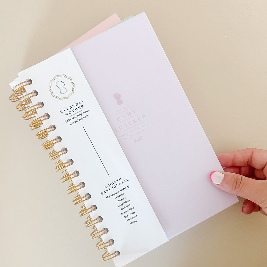 A hand touching the corner of a light purple Everyday Mother Baby Tracker book showing the ivory cover, white cover slip with gold logo, and gold spiral rings