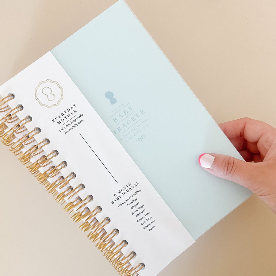 A hand touching a blue Everyday Mother Baby Tracker book showing the ivory cover, white cover slip with gold logo, and gold spiral rings