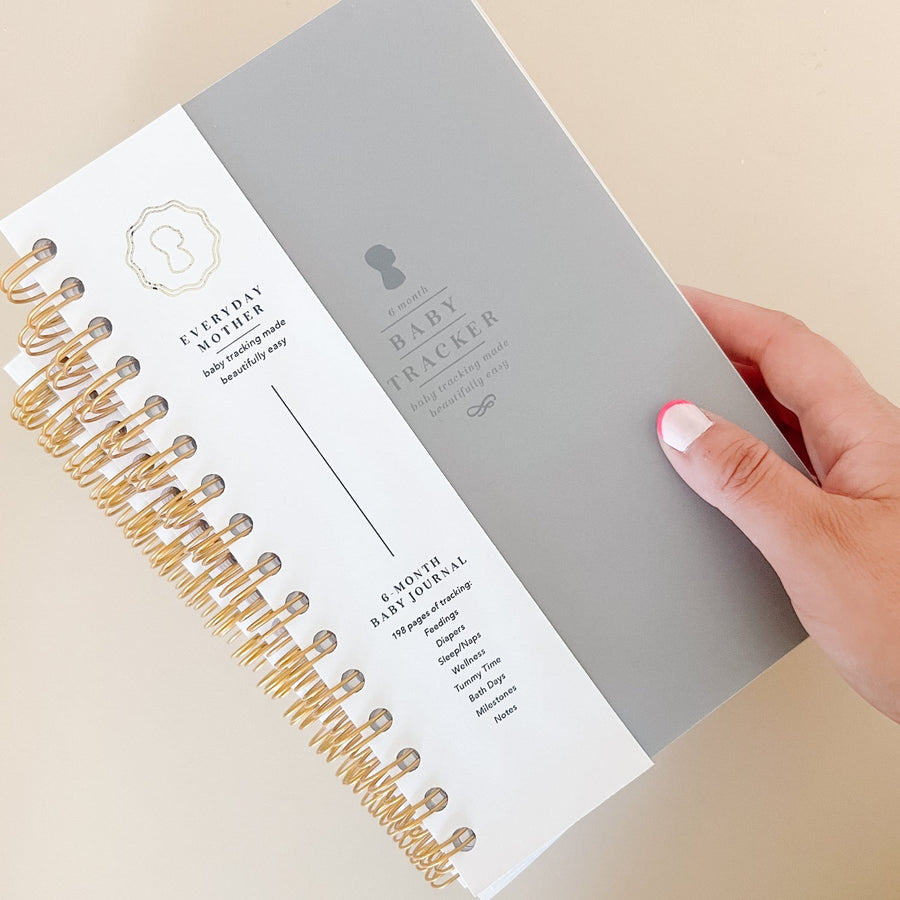 A hand touching the corner of a grey Everyday Mother Baby Tracker book showing the ivory cover, white cover slip with gold logo, and gold spiral rings