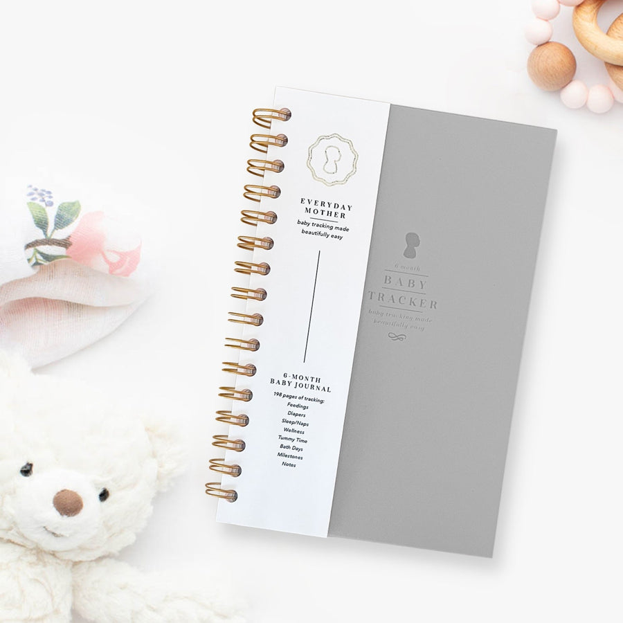 A closed grey Everyday Mother Baby Tracker book showing the ivory cover, white cover slip with gold logo, and gold spiral rings