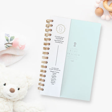 A closed light blue Everyday Mother Baby Tracker book showing the ivory cover, white cover slip with gold logo, and gold spiral rings