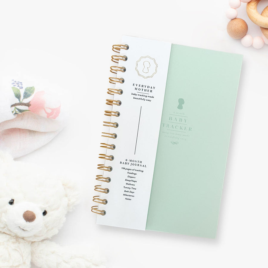 A closed mint green Everyday Mother Baby Tracker book showing the ivory cover, white cover slip with gold logo, and gold spiral rings
