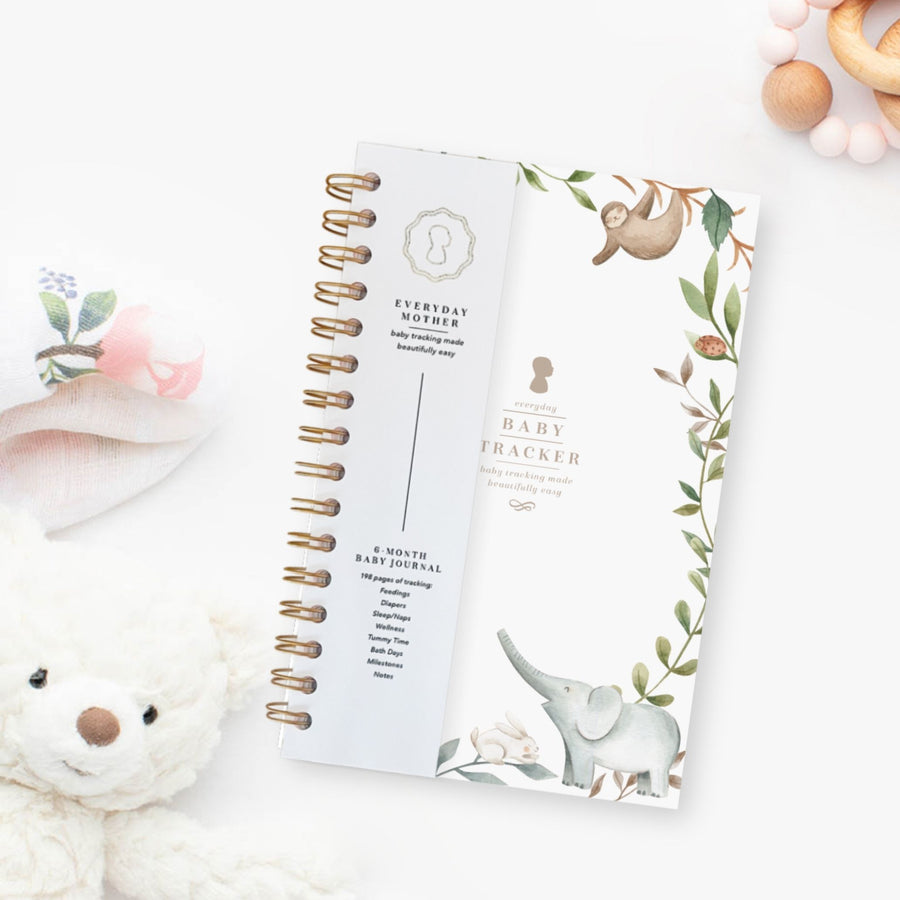 Everyday Mother baby tracker journal with gold spiral binding. Tracks 6 months of feedings, sleep, diapers, tummy time, bath days, wellness, and milestones. Background includes a plush teddy bear and a soft floral cloth.